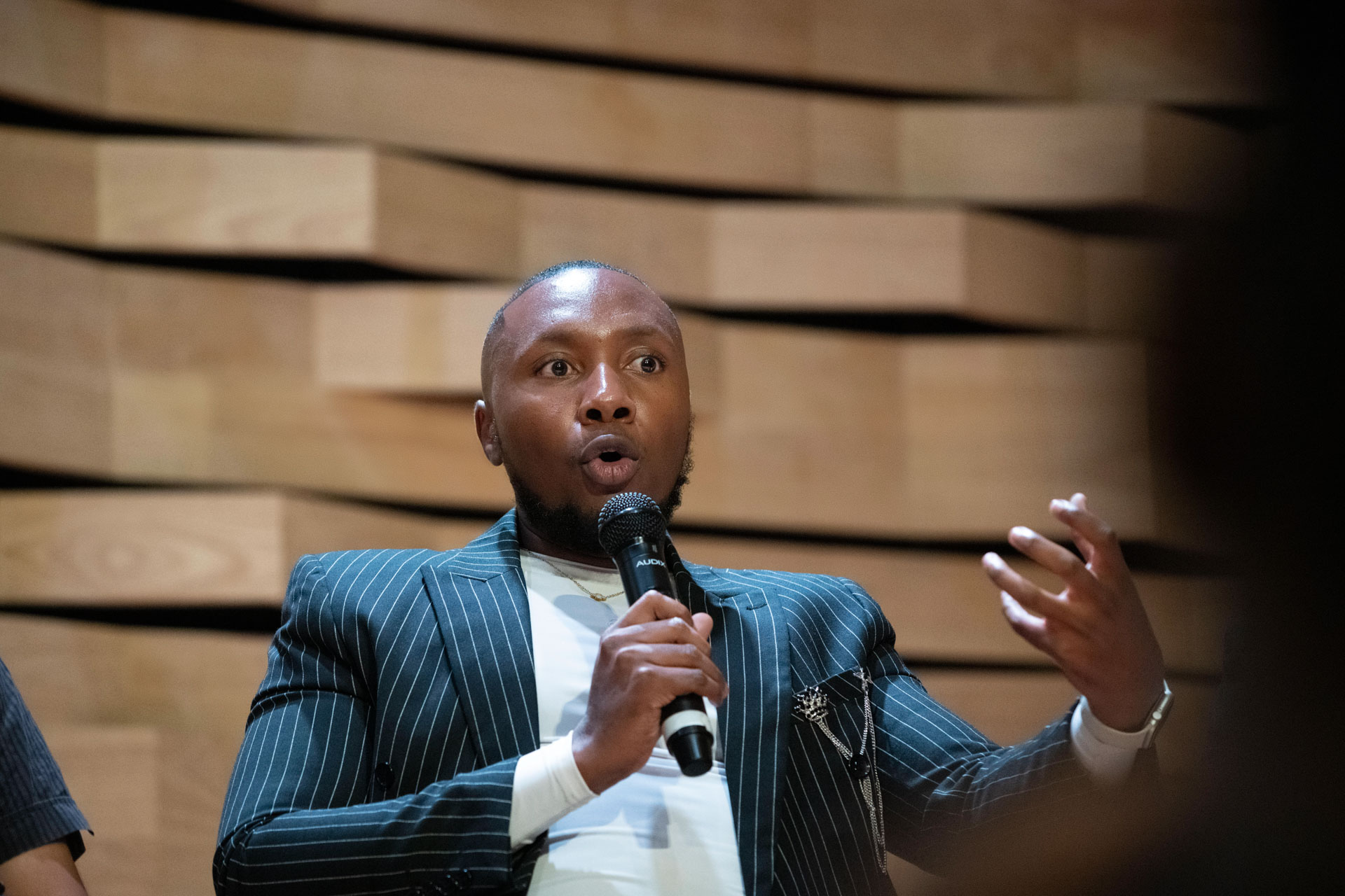 Gift Lubele speaking on stage with a microphone, pinstripe jacket, wood-slat wall. Photograph.