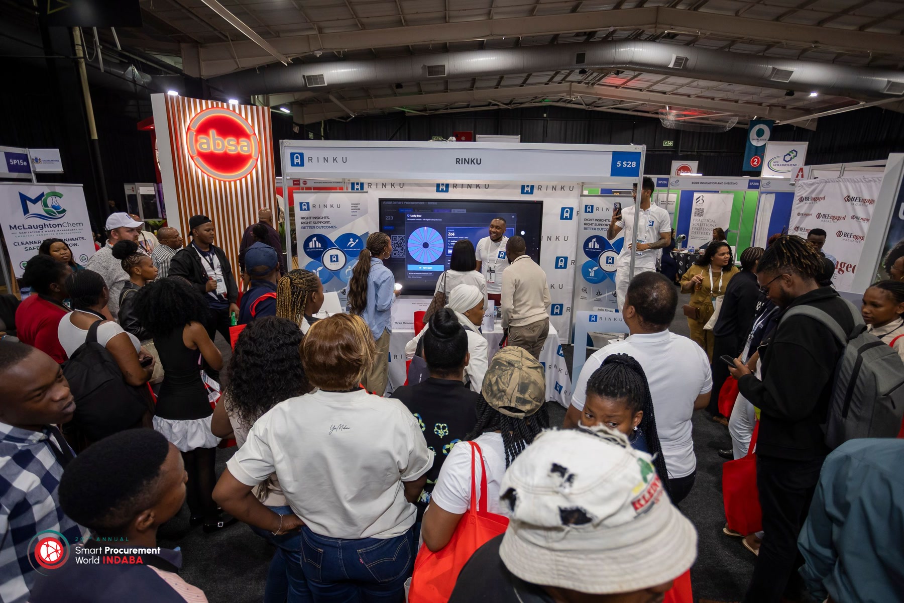 Crowd around the Rinku exhibition booth at Smart Procurement World Indaba, with a spin-wheel screen and the Absa stand nearby. Photograph.